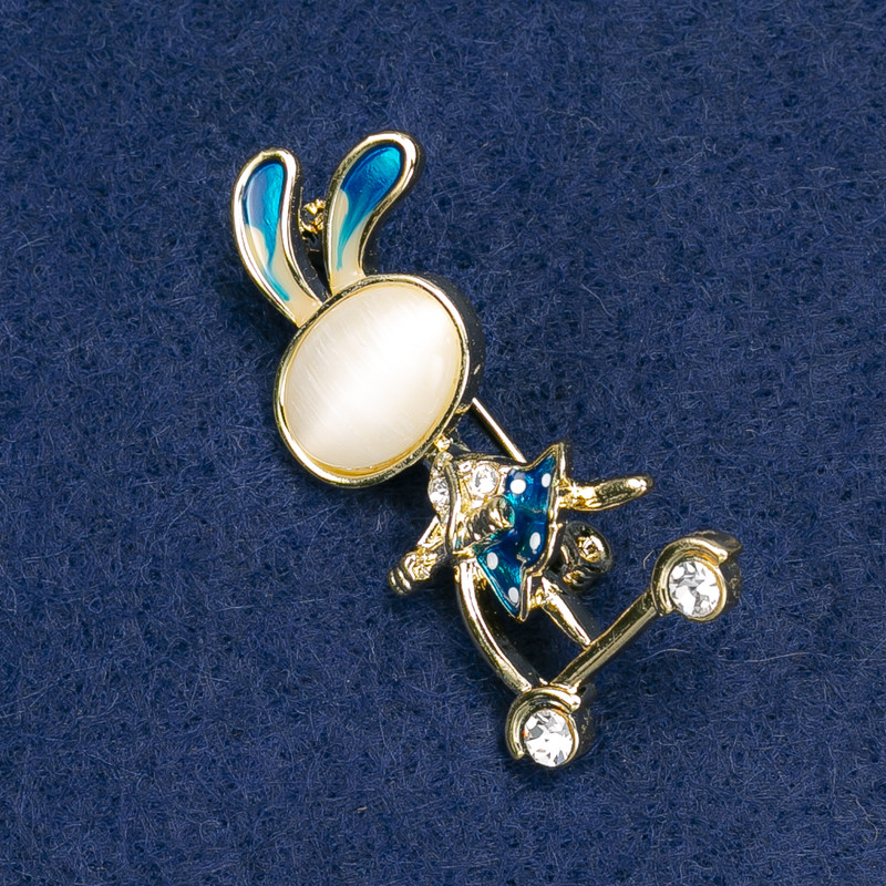 Broșă cu iepuraș pe trotinetă, cu strasuri albe, metal auriu, email albastru, cabochon piatră naturală «ochiul pisicii» alb 43×21 mm +
